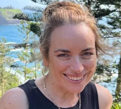 Dr. Catherine Douglass smiles at the camera. She has brown hair that is in a bun and is wearing a black short sleeve top in a beachy outdoor setting
